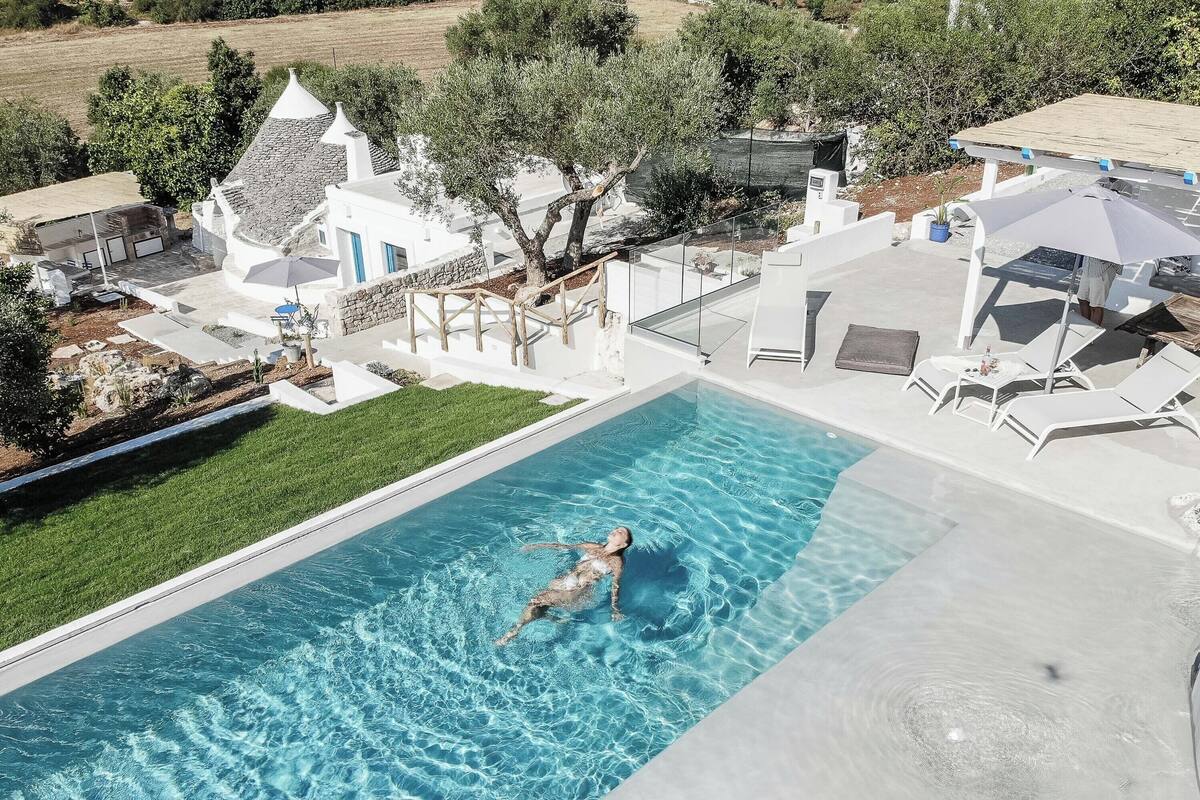 Une piscine avec une personne à l’intérieur, entourée d’une maison moderne à l’extérieur blanc et au toit en pierre.