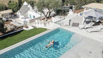 A swimming pool with a person in the water, surrounded by a modern house with a white exterior and a stone roof.