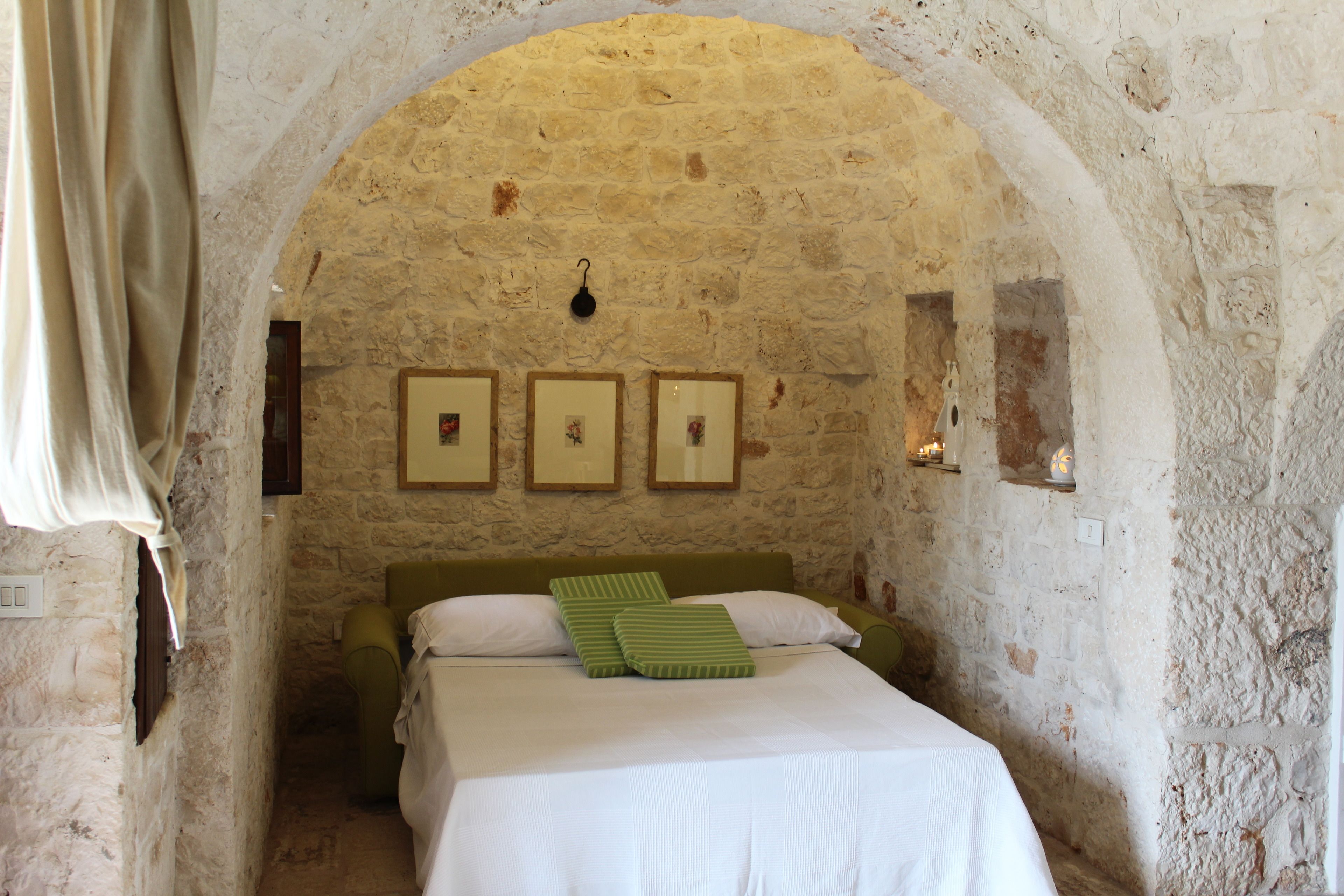 A narrow room with a stone wall, a bed with white bedding and green pillows, and three framed pictures on the wall.
