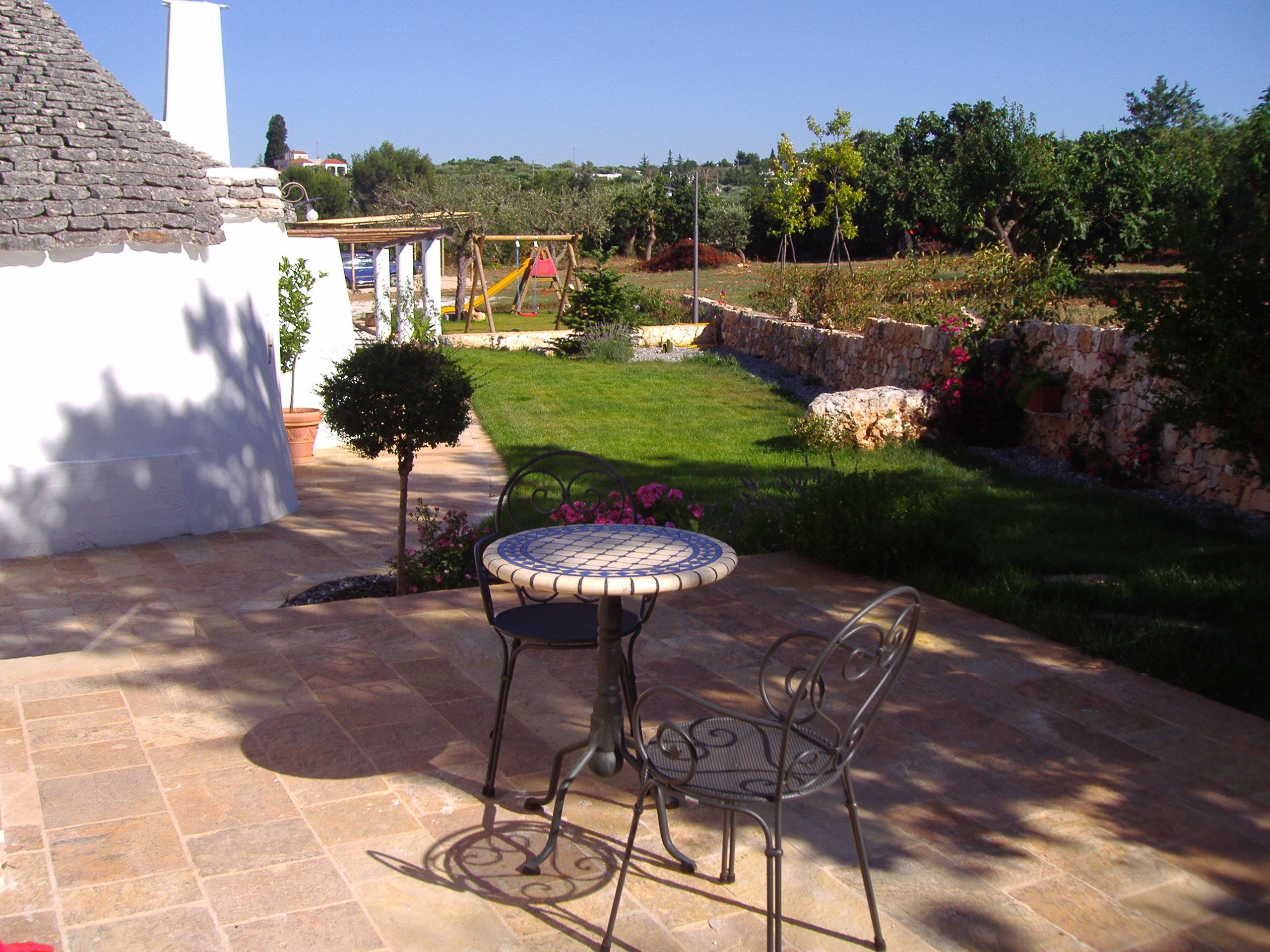 Une terrasse avec une table ronde et deux chaises, un petit arbre et un jardin avec une structure de jeux en arrière-plan.