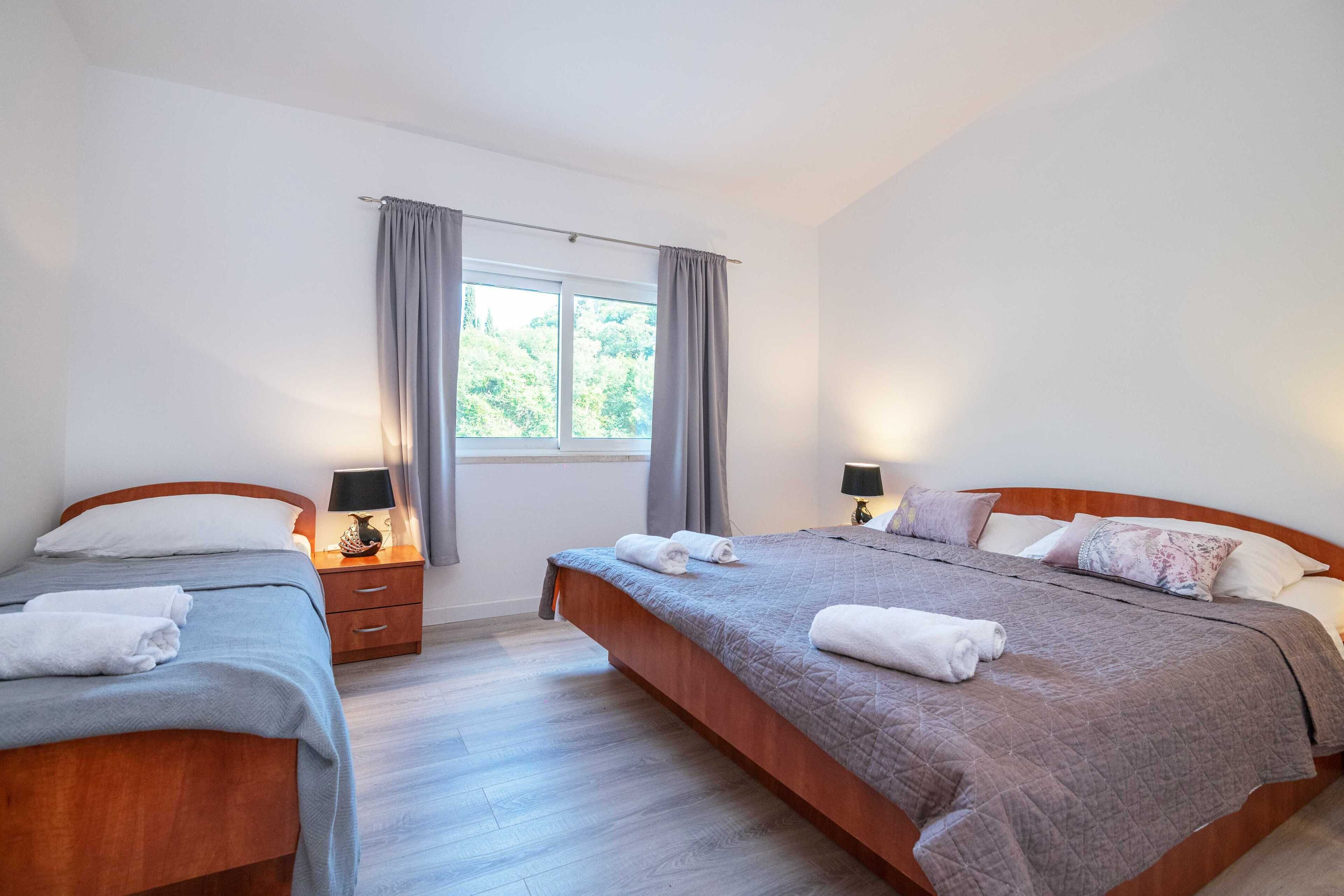 Ein Doppelzimmer mit zwei Betten, einem Fenster mit Vorhängen, einem Nachttisch mit Lampe und Blick auf Grünfläche.