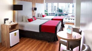 A hotel room with two beds, a small fridge, a desk, and a view of buildings.