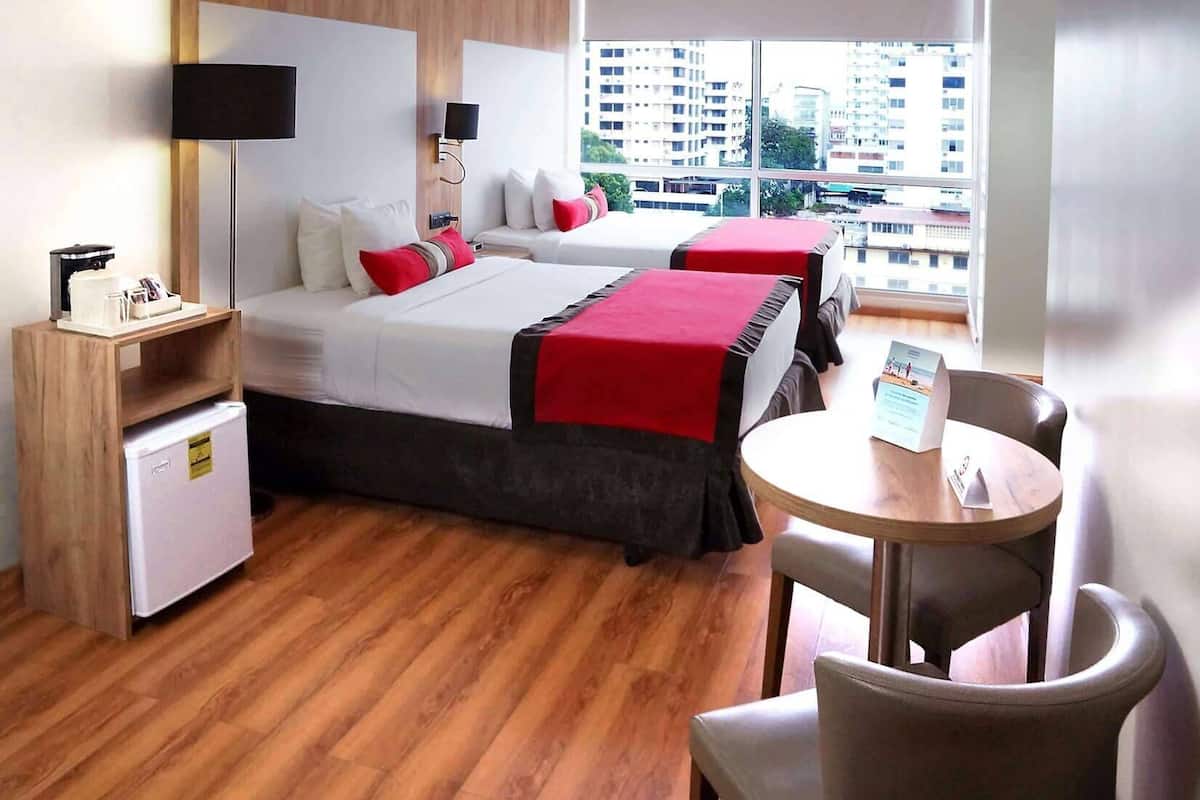 A hotel room with two beds, a small fridge, a desk, a chair, and a view of the city.