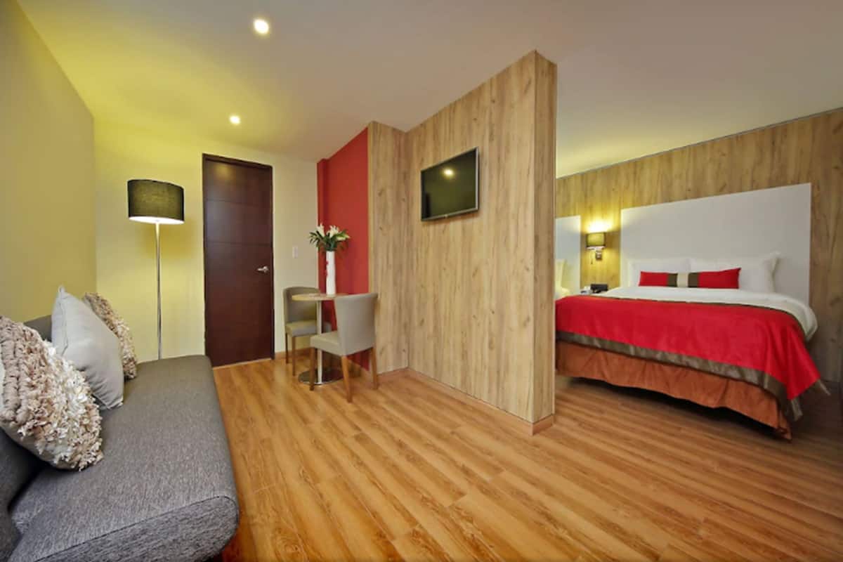 A hotel room with a wooden floor, a bed with red bedding, a grey sofa with decorative pillows, a desk with a chair, and a television mounted on the wall.