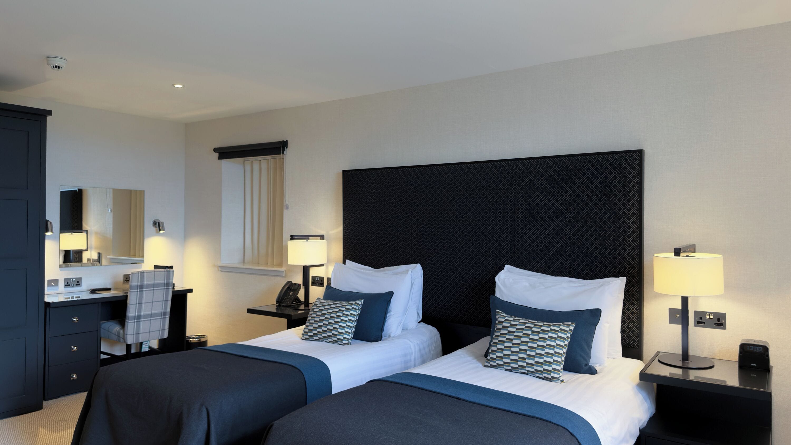 Habitación de hotel con dos camas, un escritorio con teléfono, un espejo y una mesita de noche con lámpara.