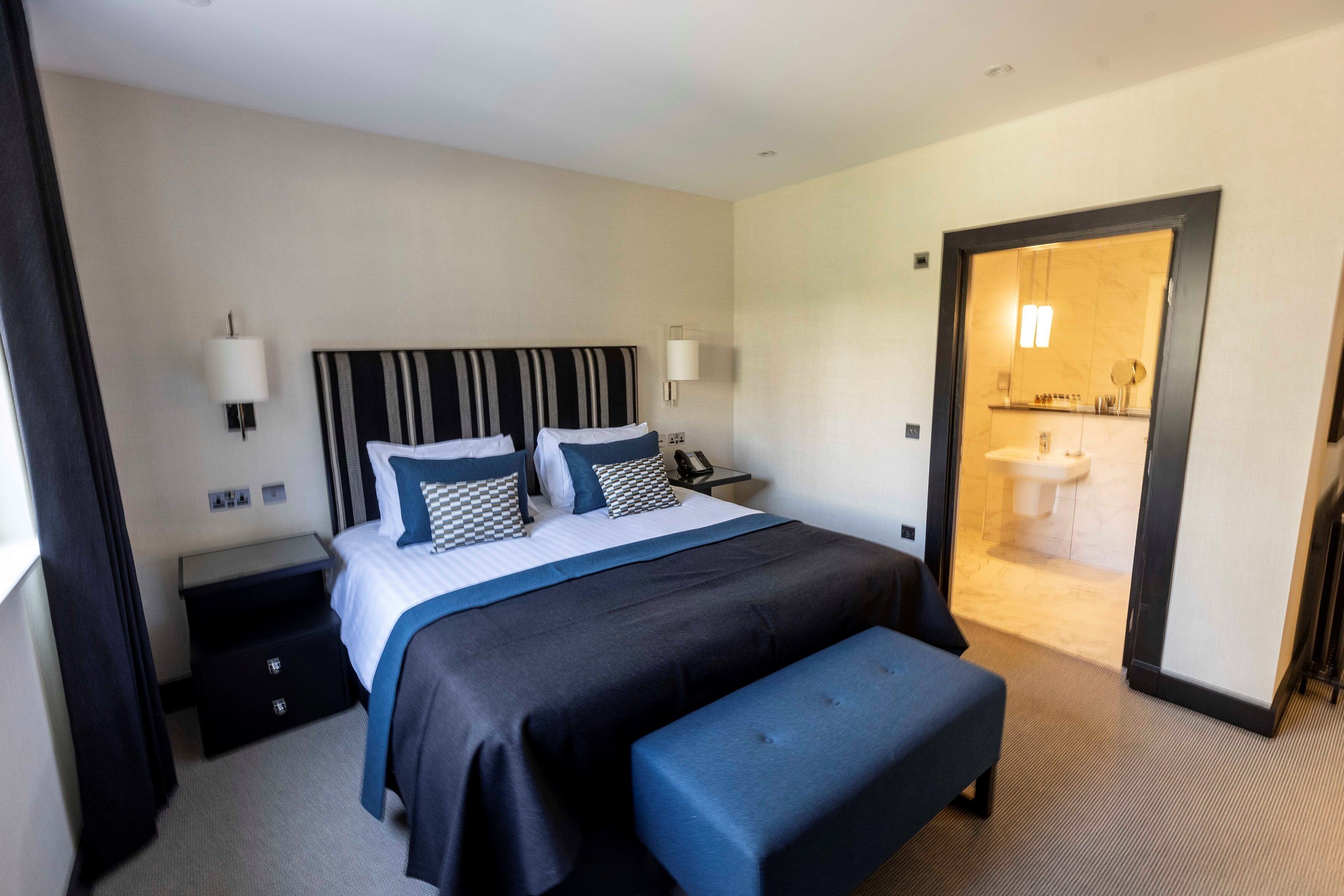 A hotel room with a large bed, bedside tables, a bench, and a bathroom visible through an open door.