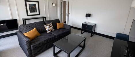 A modern hotel room with a black sofa, a glass coffee table, a desk with a computer, and a framed picture on the wall.
