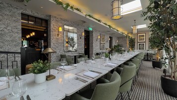 Un lungo tavolo da pranzo apparecchiato, con sedie, un'area bar e piante ornamentali.