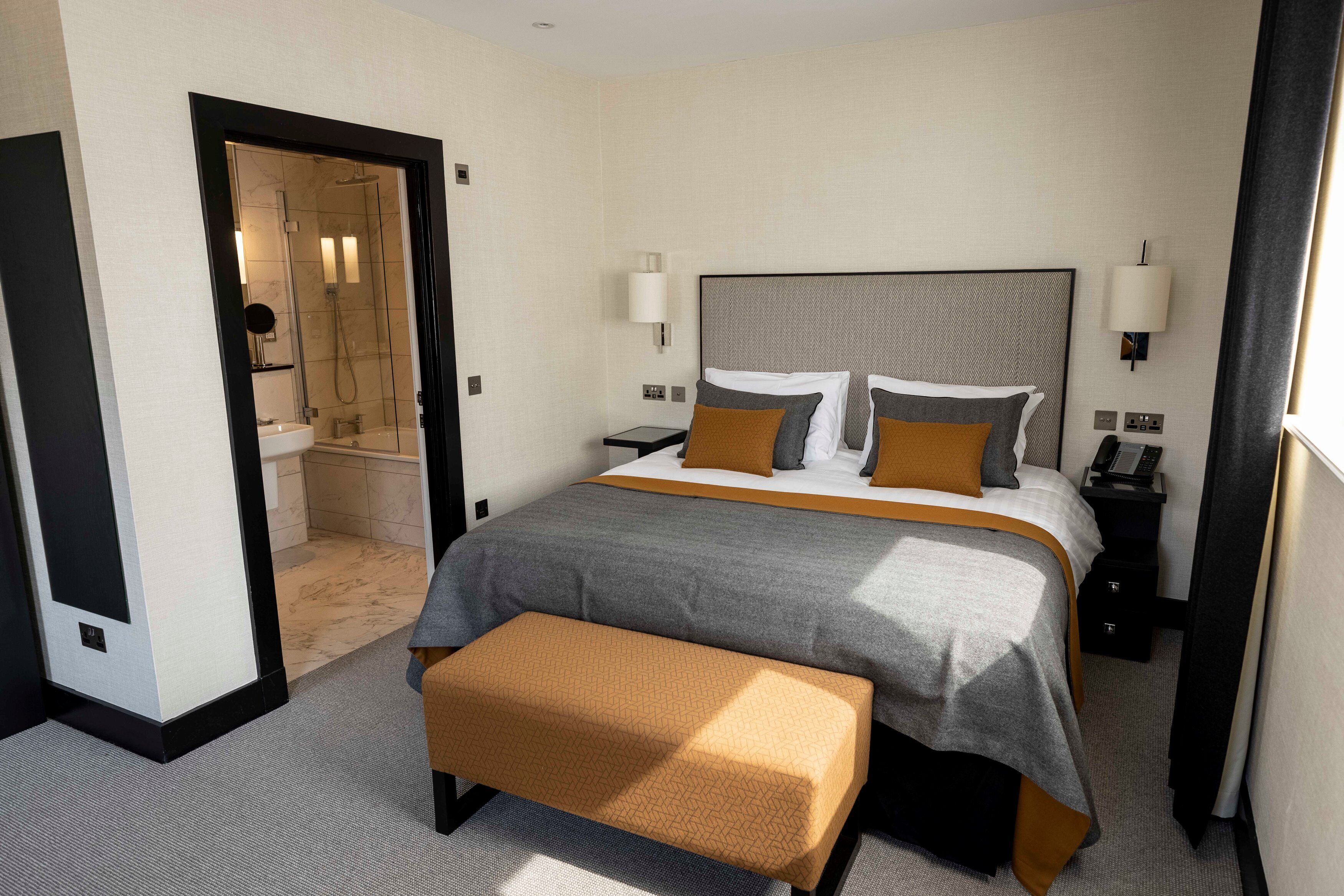 A hotel room with a large bed, bedside tables, a bench, a shower area, and a door.