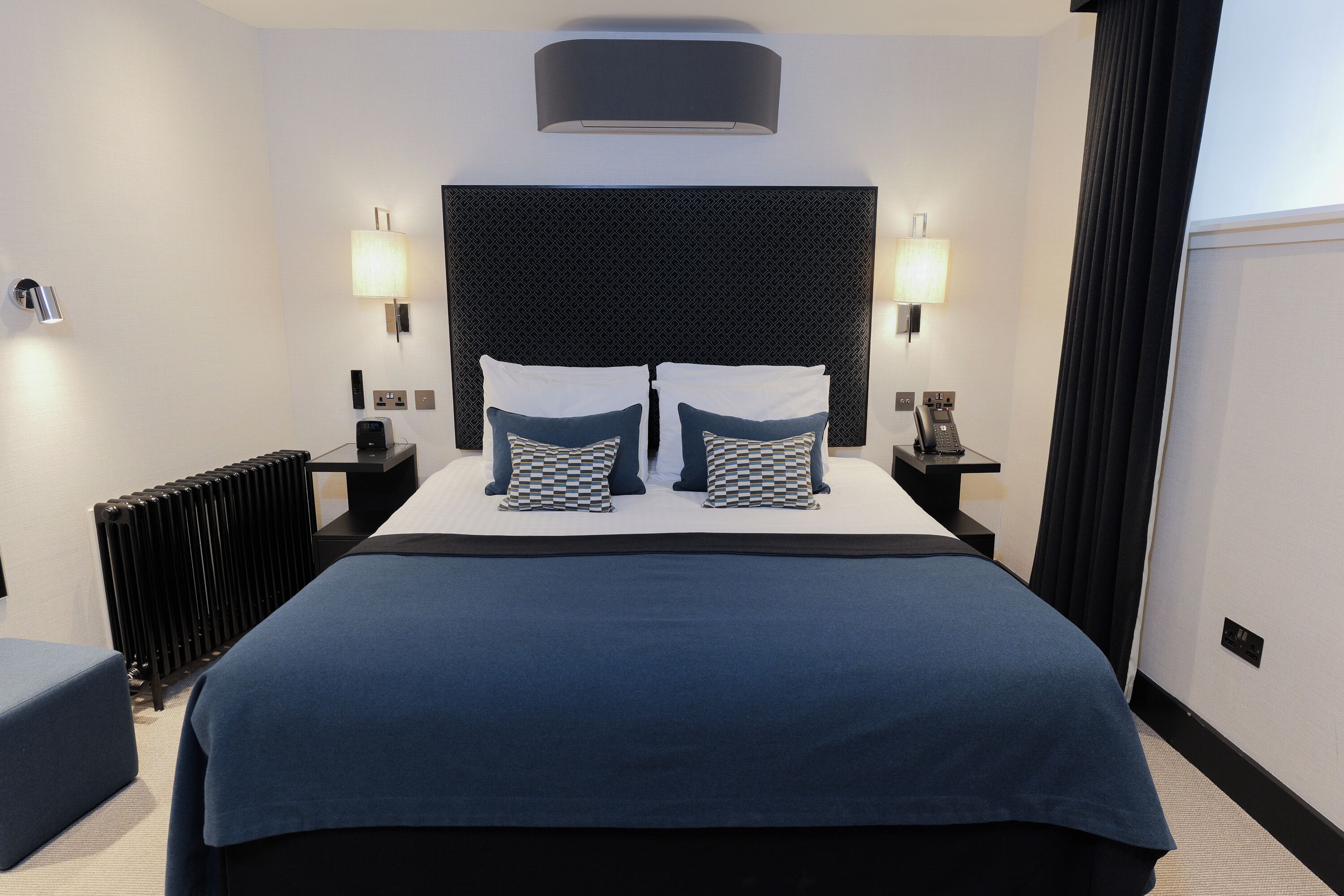 Habitación de hotel con una cama grande, dos lámparas de noche, cabecero negro y un radiador.