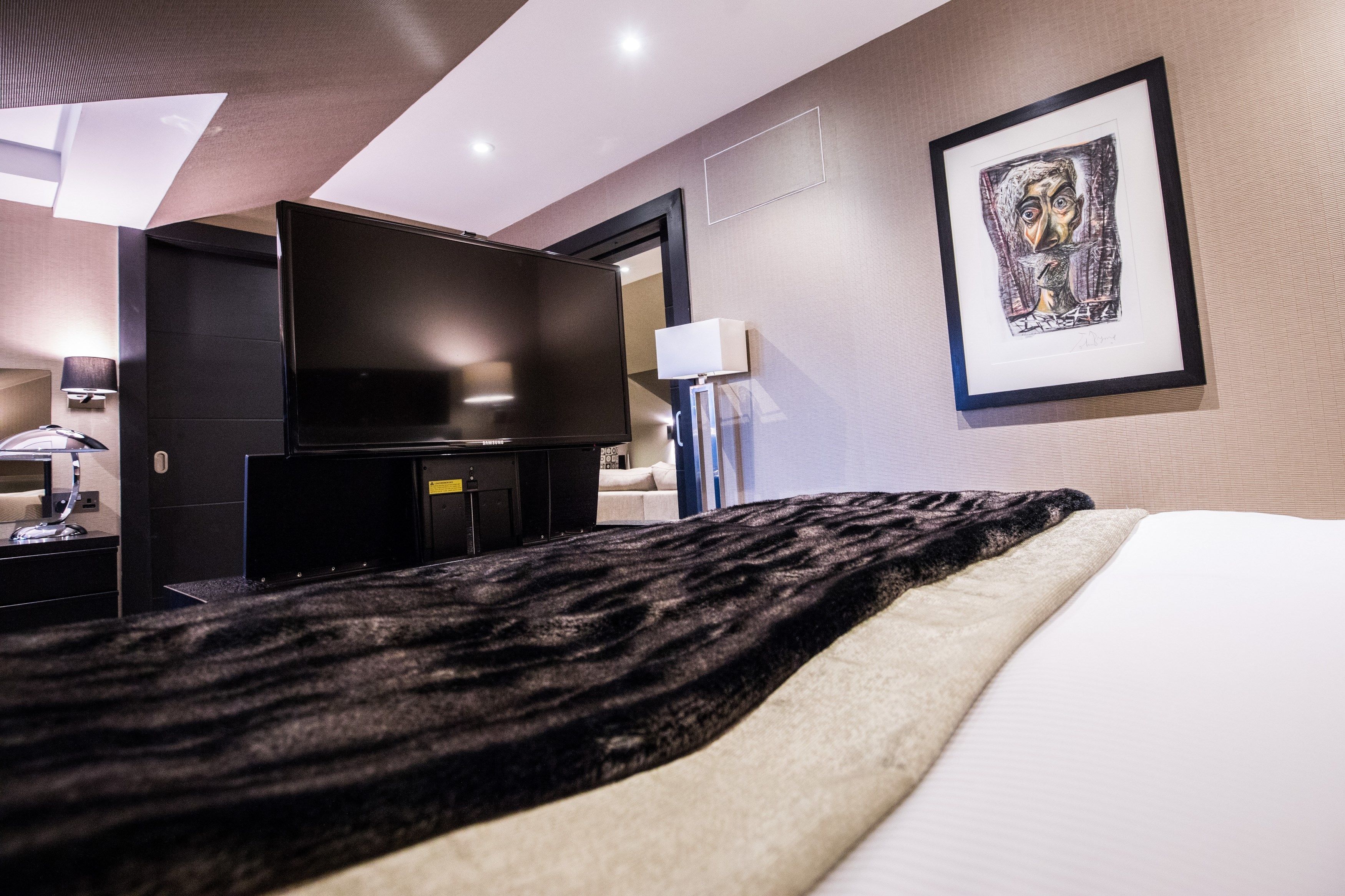 Ein modernes Hotelzimmer mit Flachbildfernseher, einem großen Bett mit schwarz-weißem Überwurf, einem gerahmten Kunstwerk an der Wand und einem Schreibtisch mit Lampe.