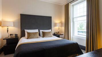 A hotel room with a large bed, two bedside tables with lamps, a window with curtains, and a view of buildings outside.