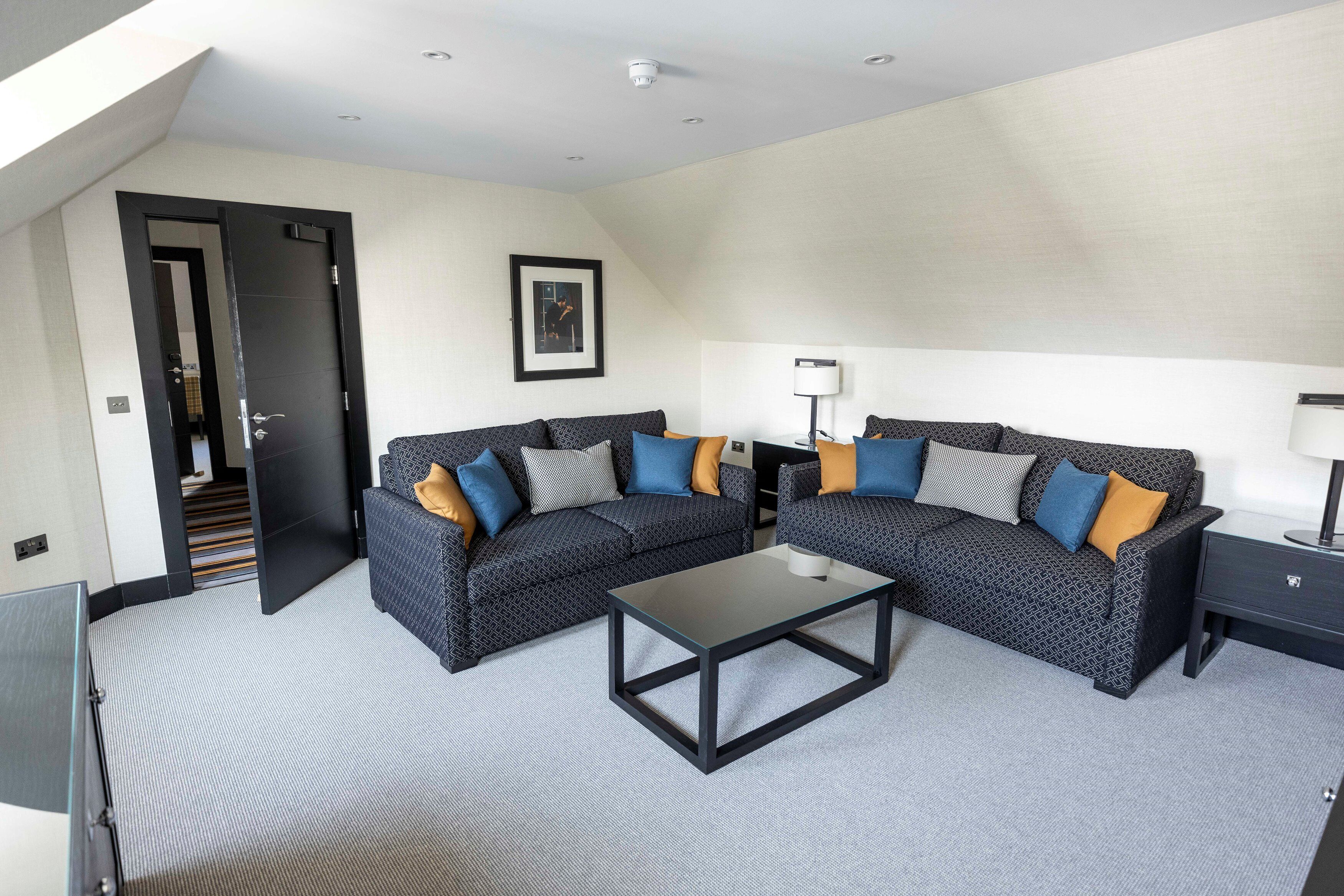A modern living room with two patterned sofas, a glass coffee table, a side table with a lamp, and a framed picture on the wall.