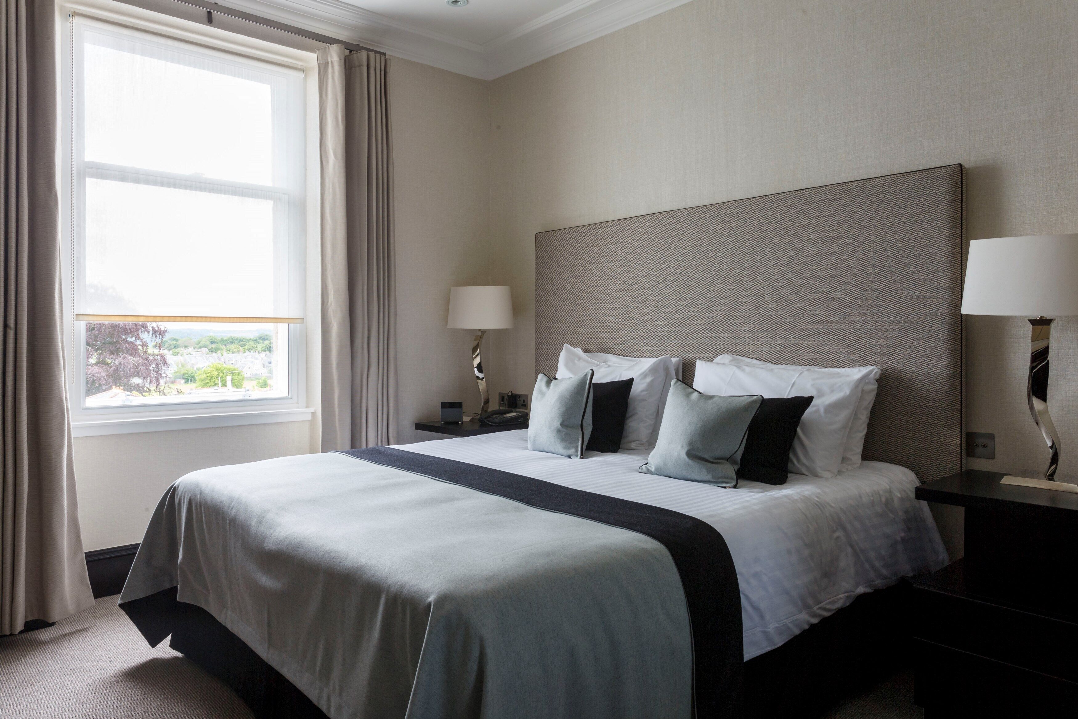 A hotel room with a large bed, two bedside tables with lamps, a window with curtains, and a view of greenery outside.