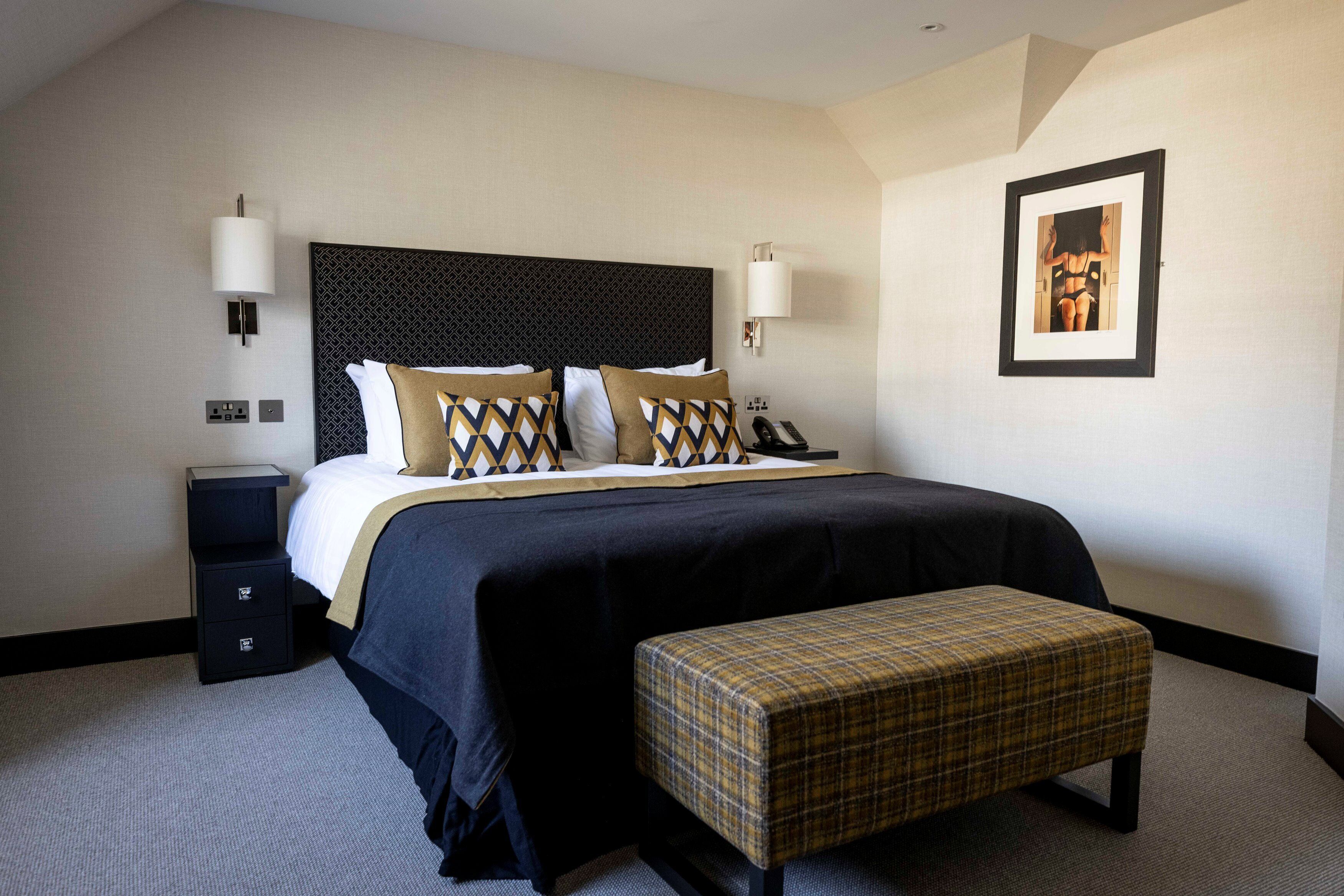A hotel room with a large bed, a bedside table, a bench, and a framed picture on the wall.