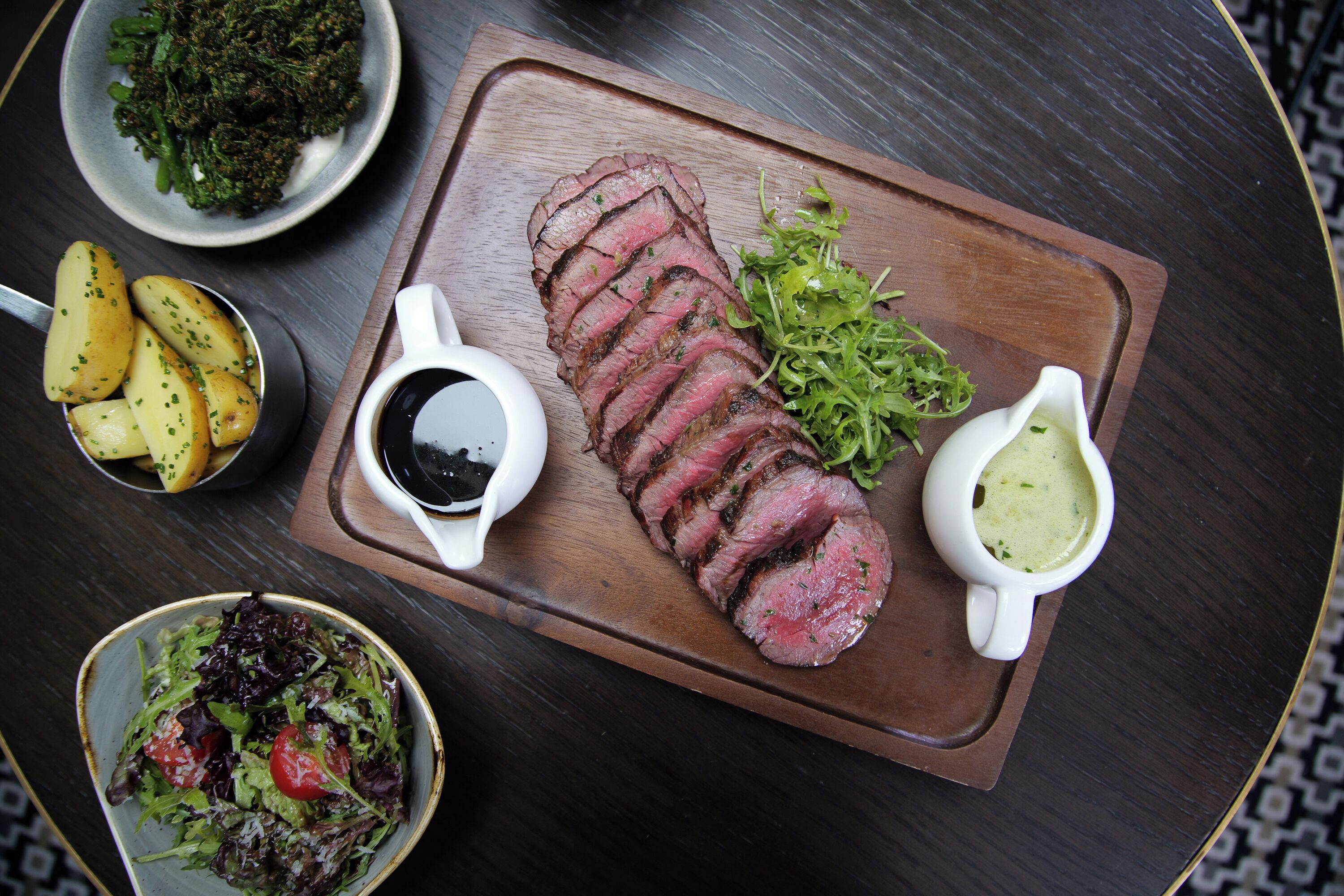 A wooden board with sliced steak, two small bowls of sauce, a plate of potatoes, and a bowl of salad.