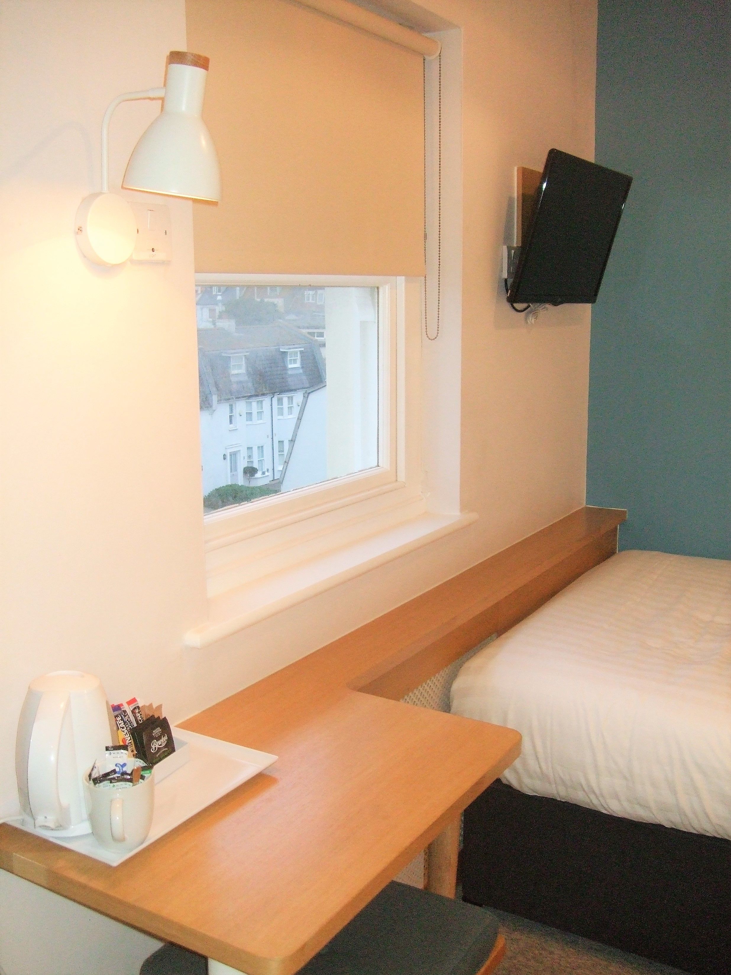 Une chambre d’hôtel avec un bureau en bois, une lampe blanche, un téléviseur à écran plat fixé au mur et une fenêtre donnant sur les bâtiments environnants.