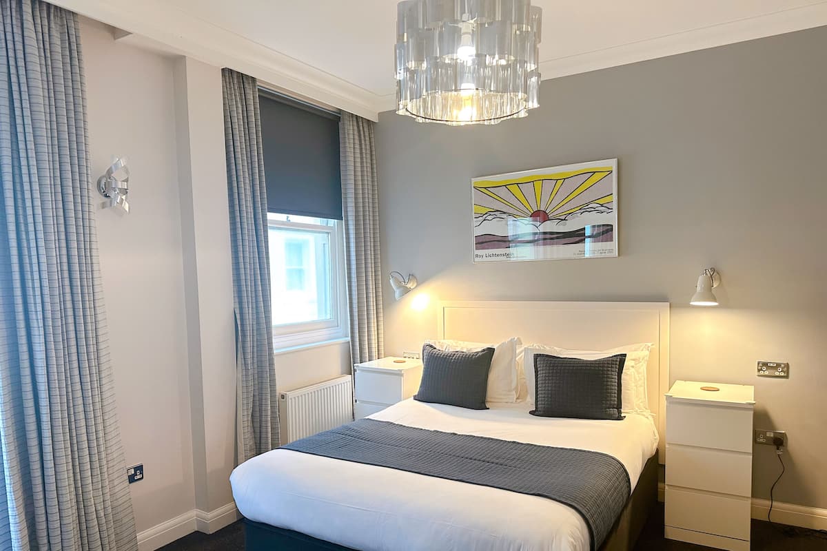 A hotel room with a bed, bedside tables, a radiator, a window with curtains, a chandelier, a wall-mounted artwork, and a desk lamp.