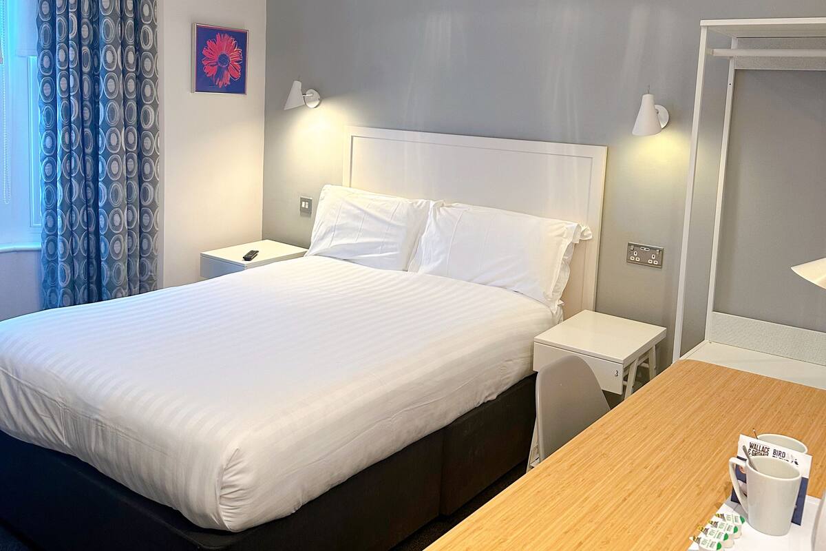 A hotel room with a bed, bedside tables, a wooden desk, a coffee cup, and a mirror.
