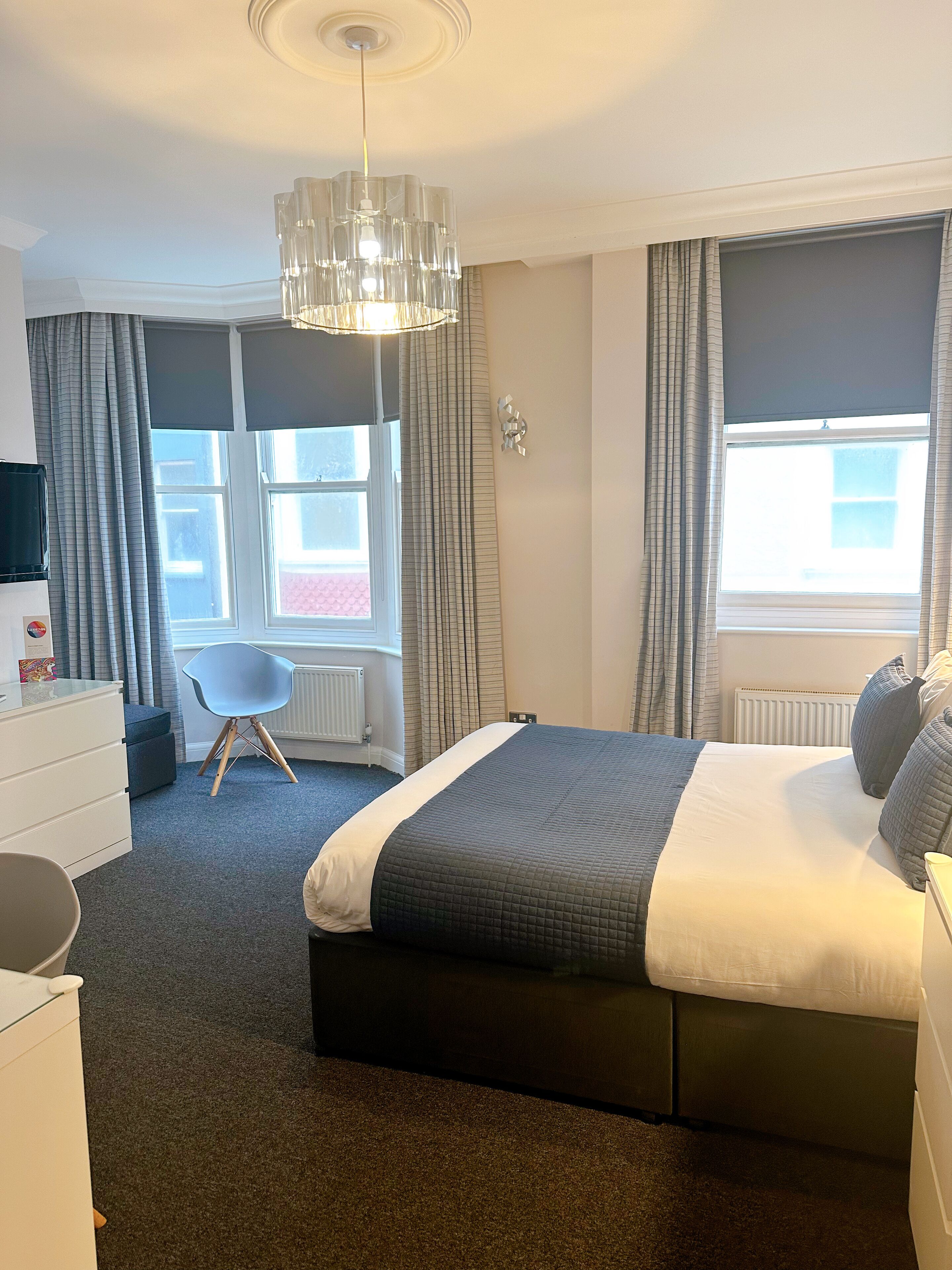 A hotel room with a bed, a dresser, a chair, and a large window with curtains.