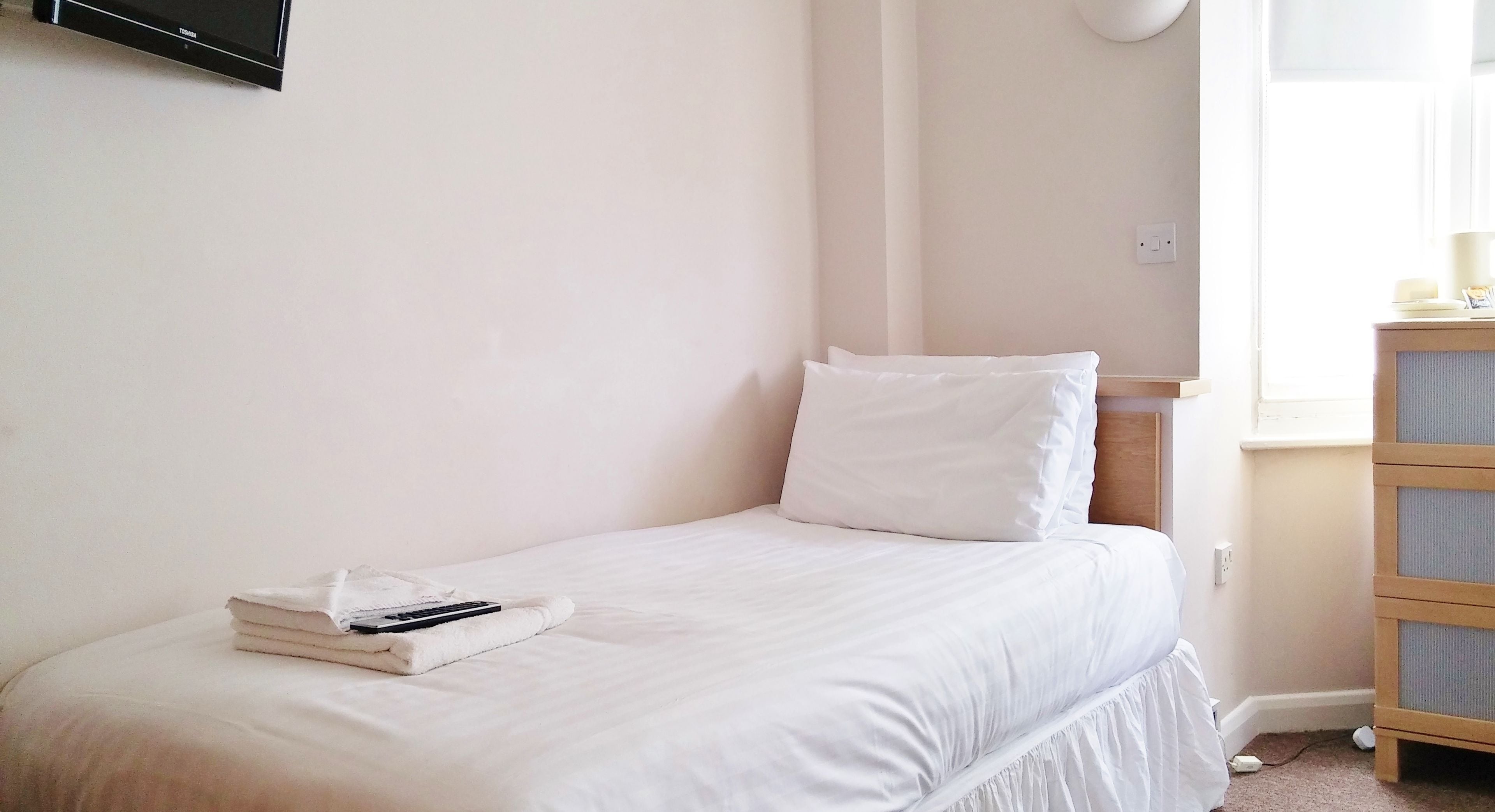 A small, neatly made bed with white bedding and pillows, a folded towel, and a remote control on the bed.