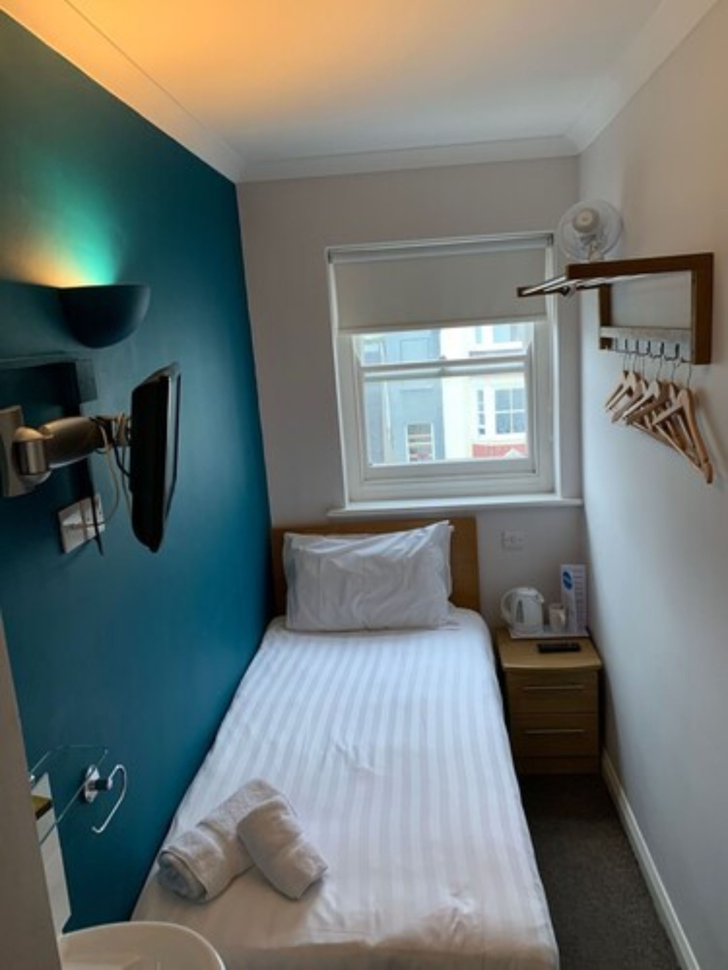 A small, neatly arranged hotel room with a single bed, a bedside table with a lamp, a window with a view of buildings, and a wall-mounted shelf with a mirror and hooks.
