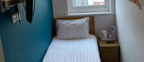 A small, neatly arranged hotel room with a single bed, a bedside table with a lamp, a window with a view of buildings, and a wall-mounted shelf with a mirror and hooks.