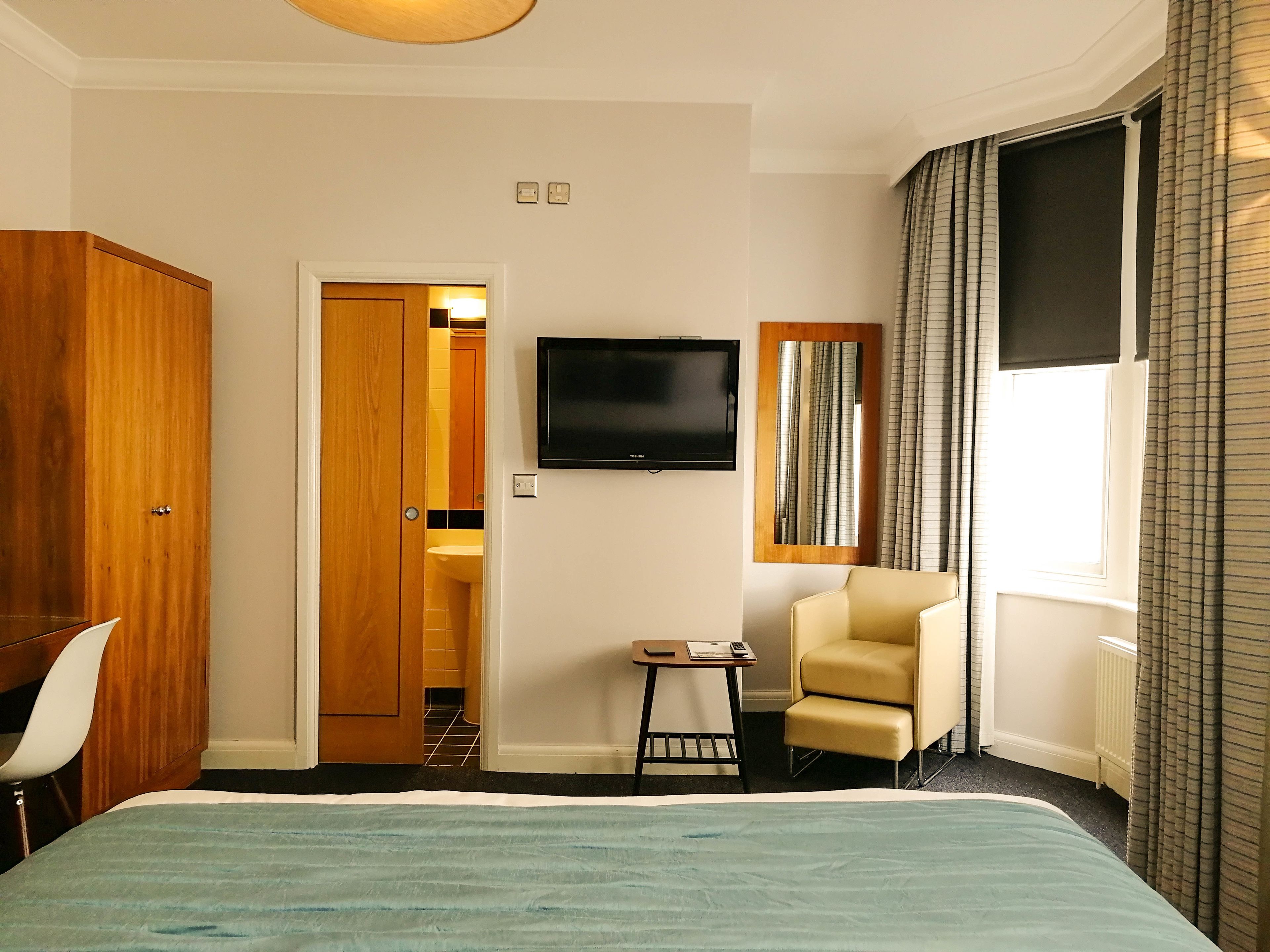 A hotel room with a bed, a television, a small table with a chair, and a wardrobe.