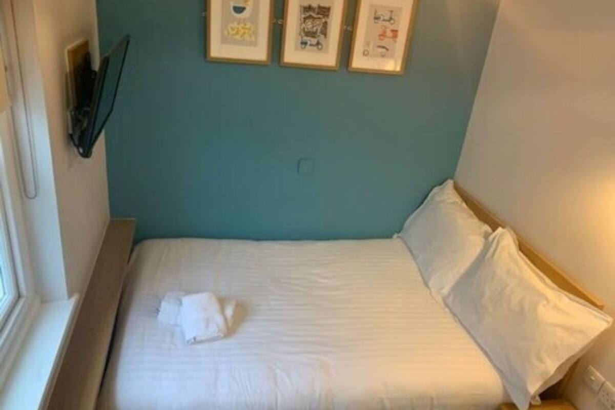 A small, neatly made bed with white linens and pillows, a window on the left, and three framed pictures on the wall above the bed.