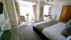 Quarto de hotel com uma cama grande, uma escrivaninha, uma cadeira e uma pequena mesa. Há uma janela com cortinas e vista para o exterior.