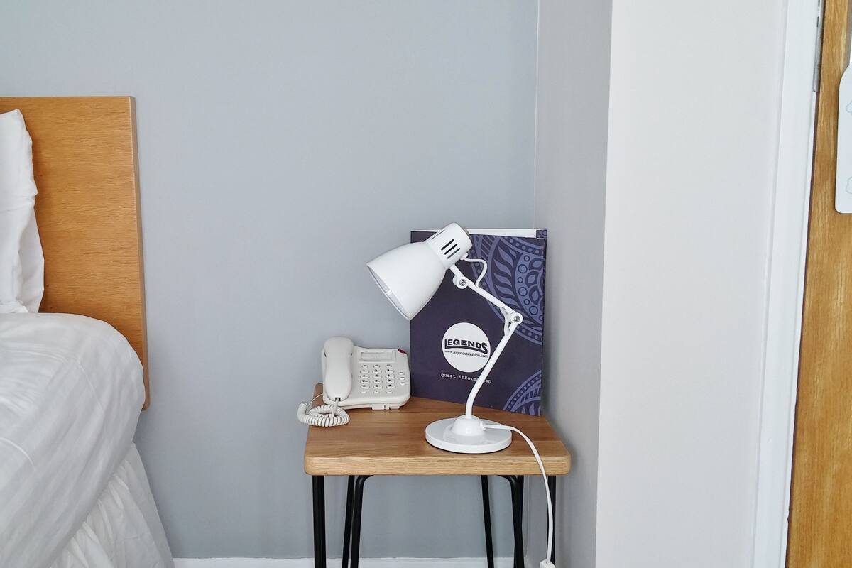 A neatly made bed with white linens, a bedside table with a white lamp, a telephone, and a framed poster on the wall.