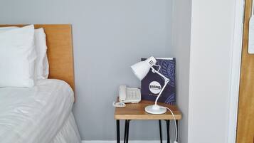 Uma cama bem arrumada com lençóis brancos, uma mesinha de cabeceira com abajur branco, um telefone e um cartaz emoldurado na parede.