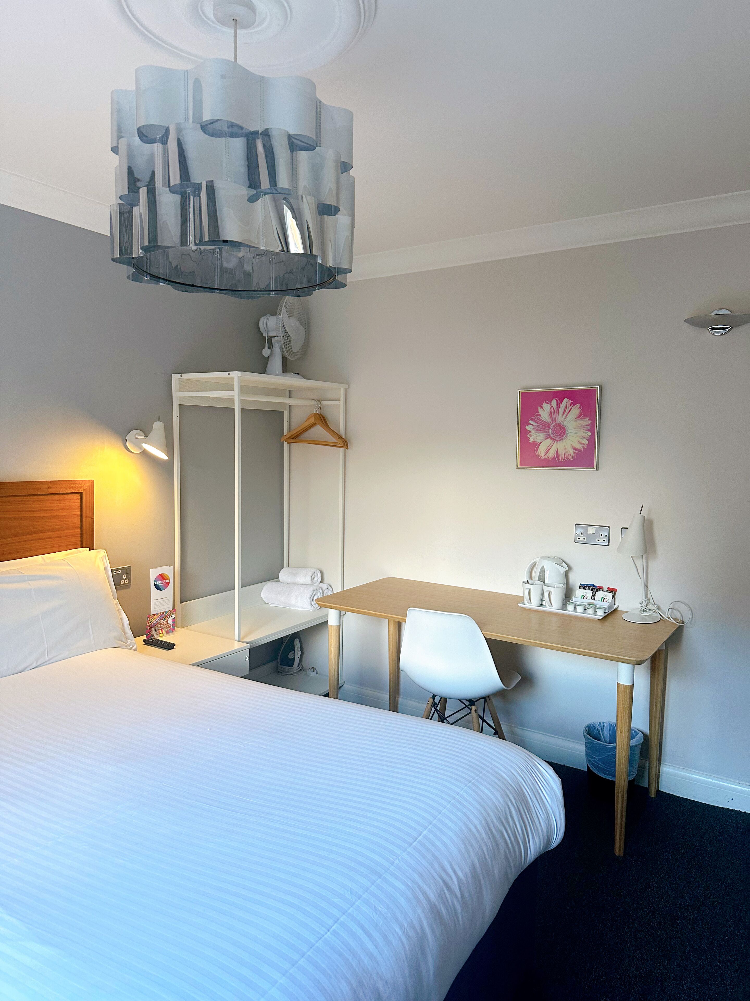 A hotel room with a bed, a desk, a chair, a wardrobe, a mirror, a hanging light fixture, a picture on the wall, and a bedside table with a lamp and a remote control.