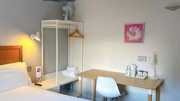 A hotel room with a bed, a desk, a chair, a wardrobe, a mirror, a hanging light fixture, a picture on the wall, and a bedside table with a lamp and a remote control.
