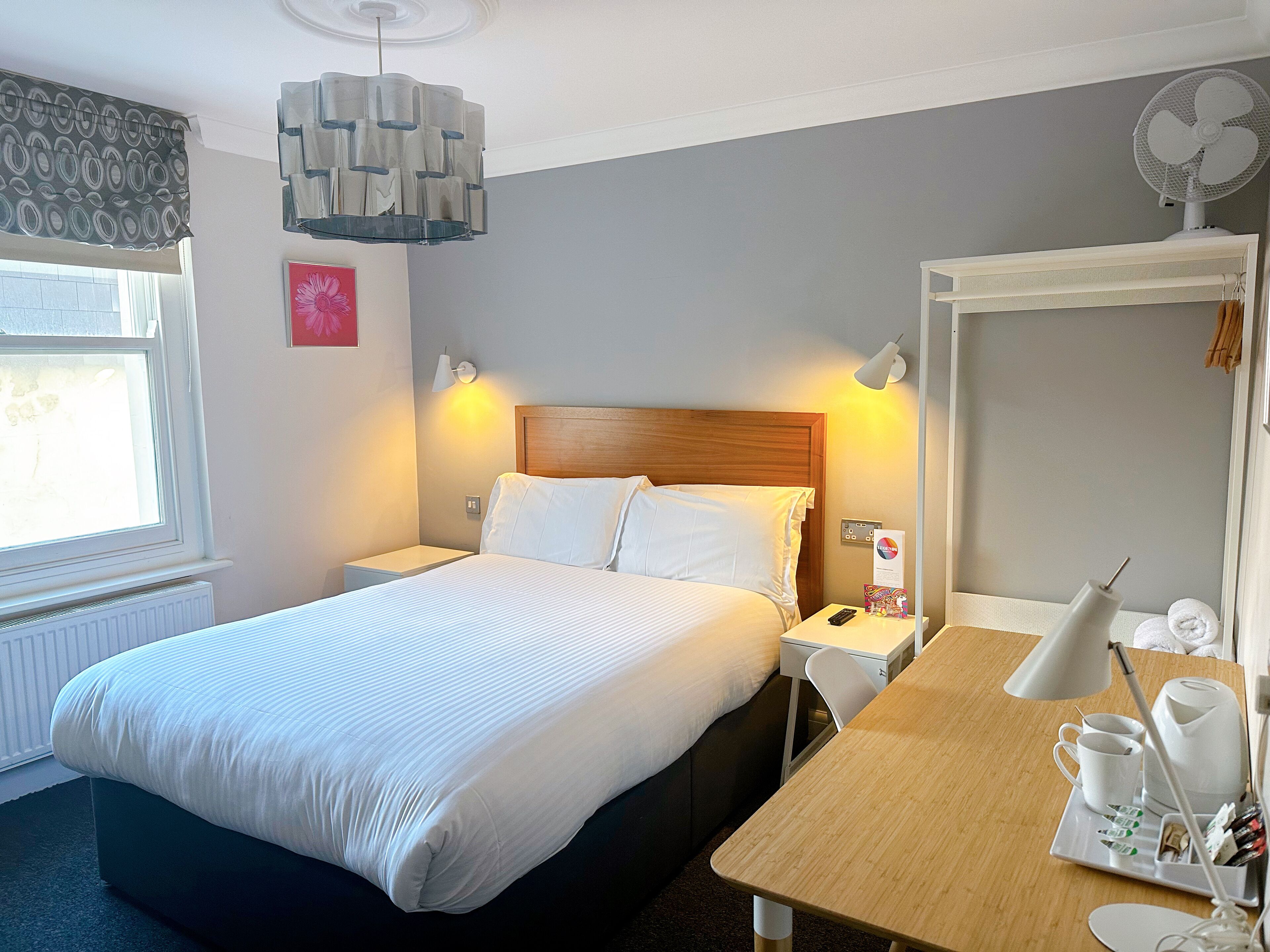 A hotel room with a large bed, bedside tables, a desk with a chair, a lamp, a fan, and a window with curtains.