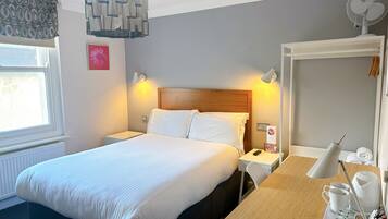 A hotel room with a large bed, bedside tables, a desk with a chair, a lamp, a fan, and a window with curtains.