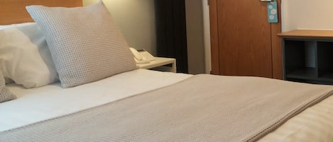 A neatly made bed with a white comforter and a beige blanket, a white pillow, a bedside table with a white telephone, and a wall-mounted lamp.