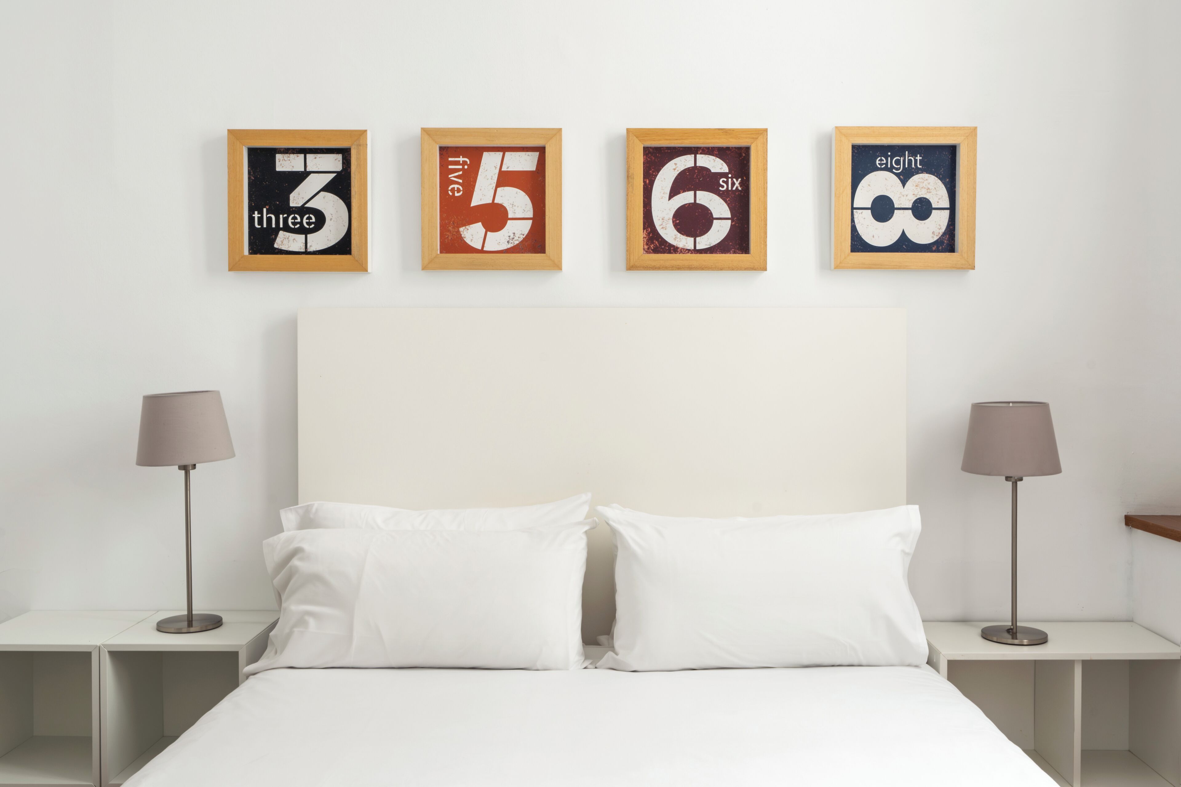 A bedroom with a bed, white pillows, a headboard, and four framed numbers on the wall.
