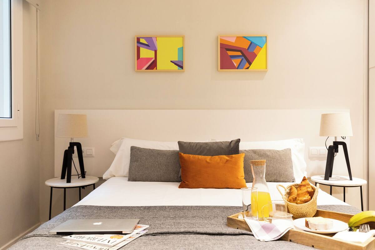 Habitación de hotel con cama, dos cuadros en la pared, una mesita de noche con lámpara, una bandeja con artículos de desayuno y un ordenador portátil sobre la cama.