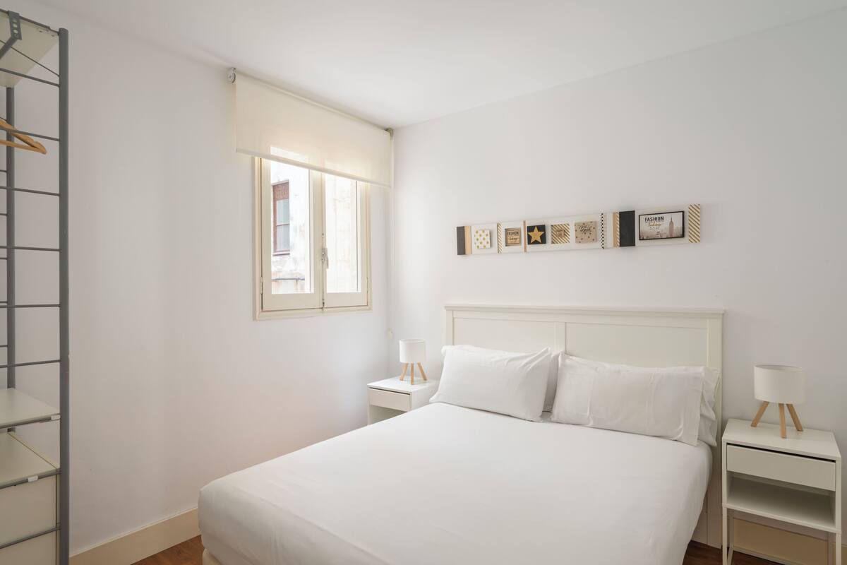 Un dormitorio minimalista con cama, mesitas de noche, una ventana y adornos en la pared.