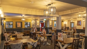 A restaurant with multiple dining tables and chairs, some with patterned cushions. There are hanging light fixtures and framed pictures on the walls. - Charnwood Arms by Greene King Inns (Coalville)