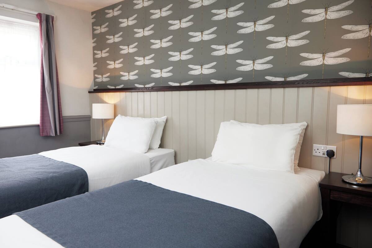 Une chambre d’hôtel avec deux lits, chacun recouvert d’une courtepointe blanche et bleue, une fenêtre avec des rideaux et un mur orné d’un motif de papillons.