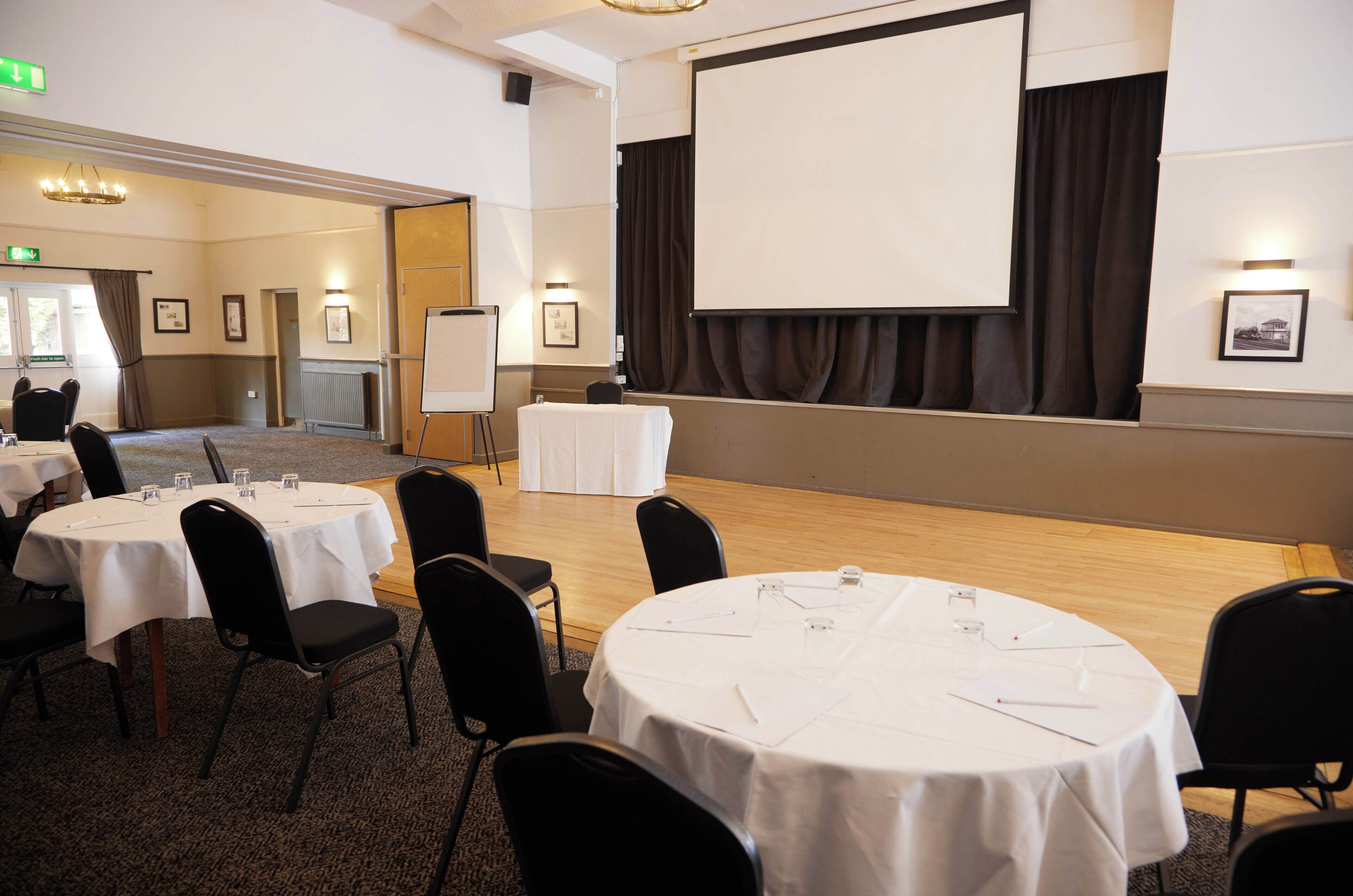 A conference room with round tables, black chairs, a whiteboard, and a projector screen.