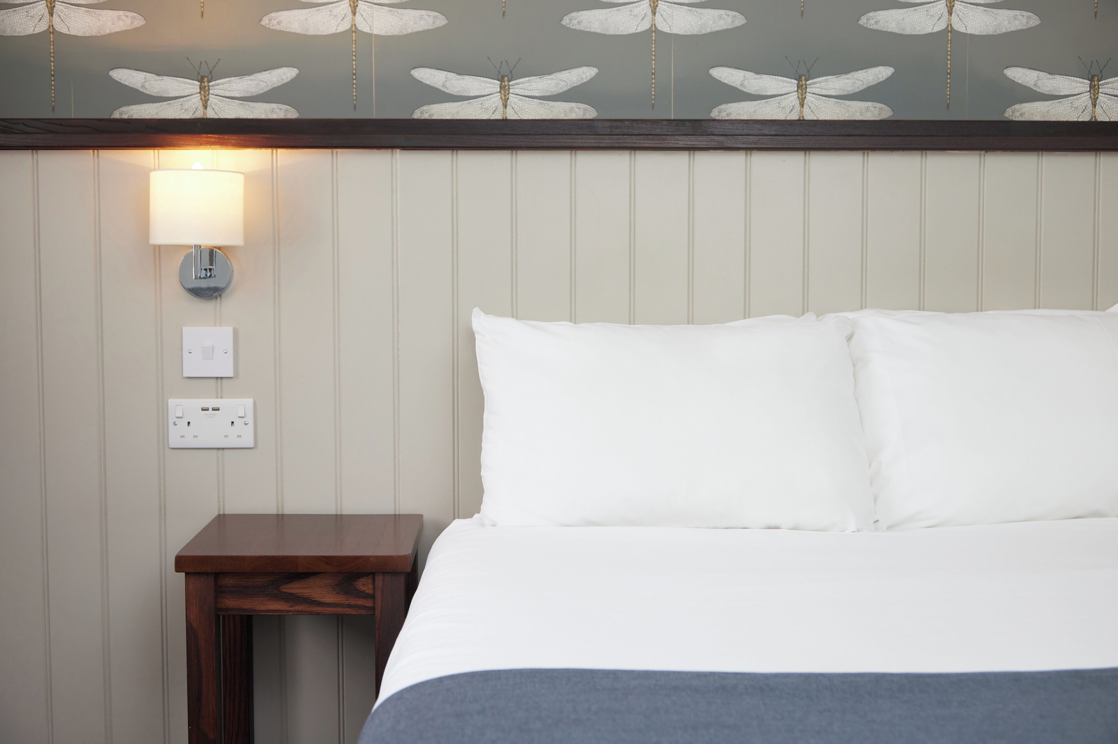 A hotel room with a bed, a nightstand, a wall-mounted lamp, and a power outlet.
