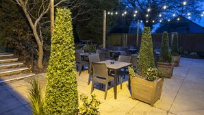 An outdoor patio area with tables and chairs, surrounded by greenery and trees. - Charnwood Arms by Greene King Inns (Coalville)