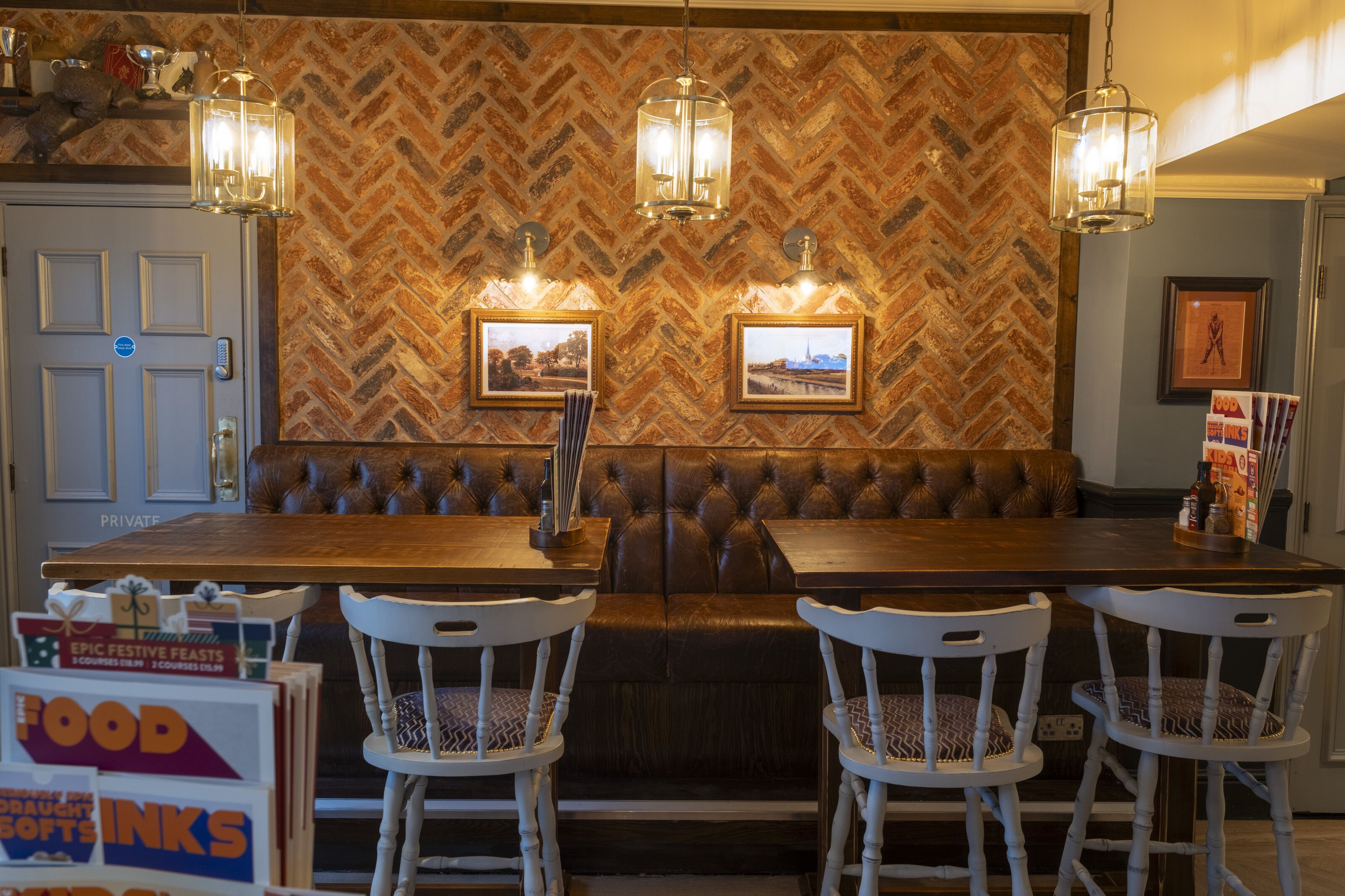 A restaurant with a herringbone-patterned brick wall, leather booth seating, and white chairs.
