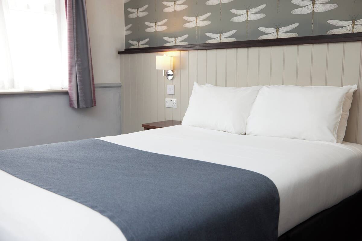 Un lit bien fait, avec du linge de lit blanc et un plaid bleu, une lampe fixée au mur et une fenêtre avec des rideaux.