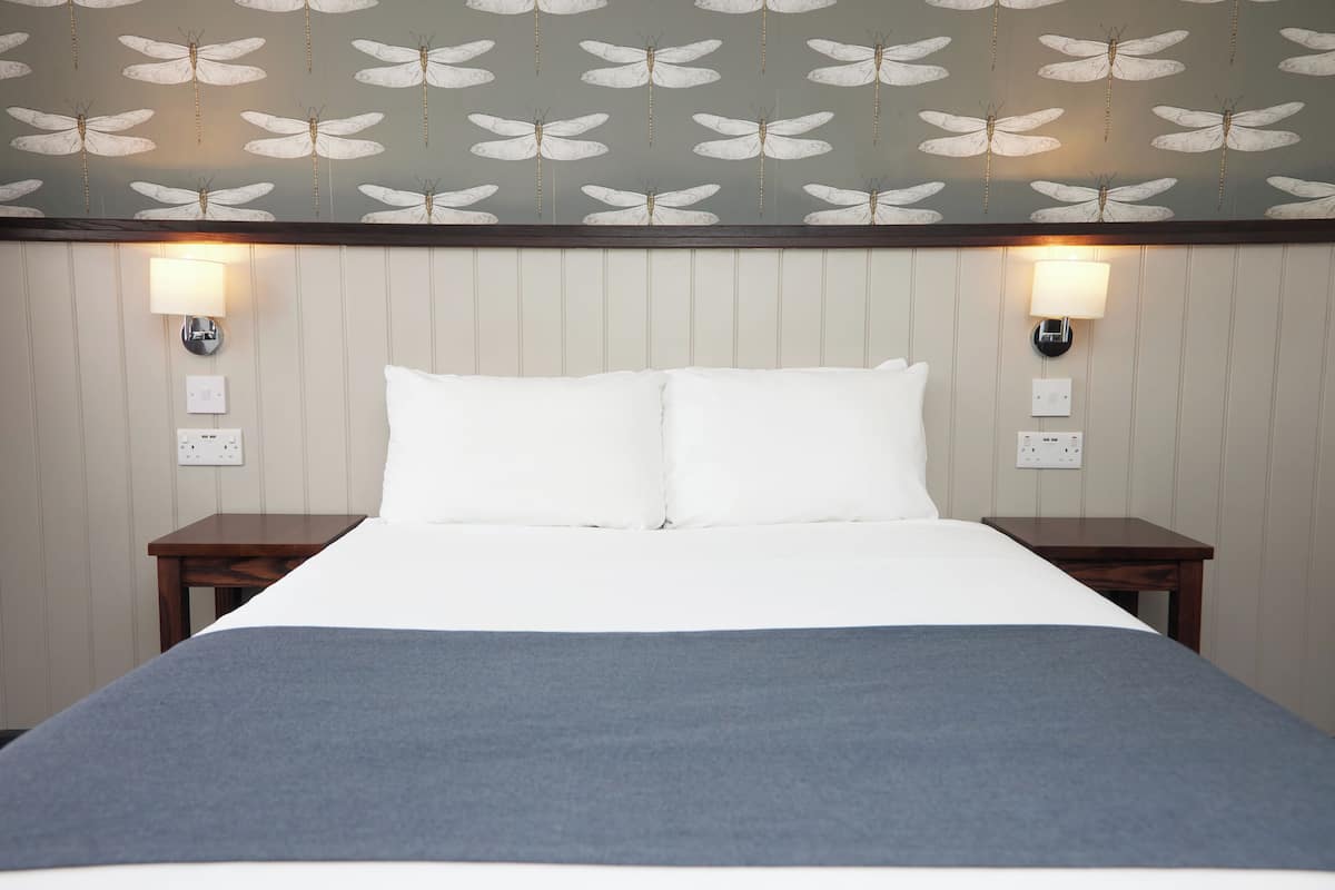 Un lit bien fait, recouvert d’une couverture grise, avec deux tables de chevet et des lampes fixées au mur.