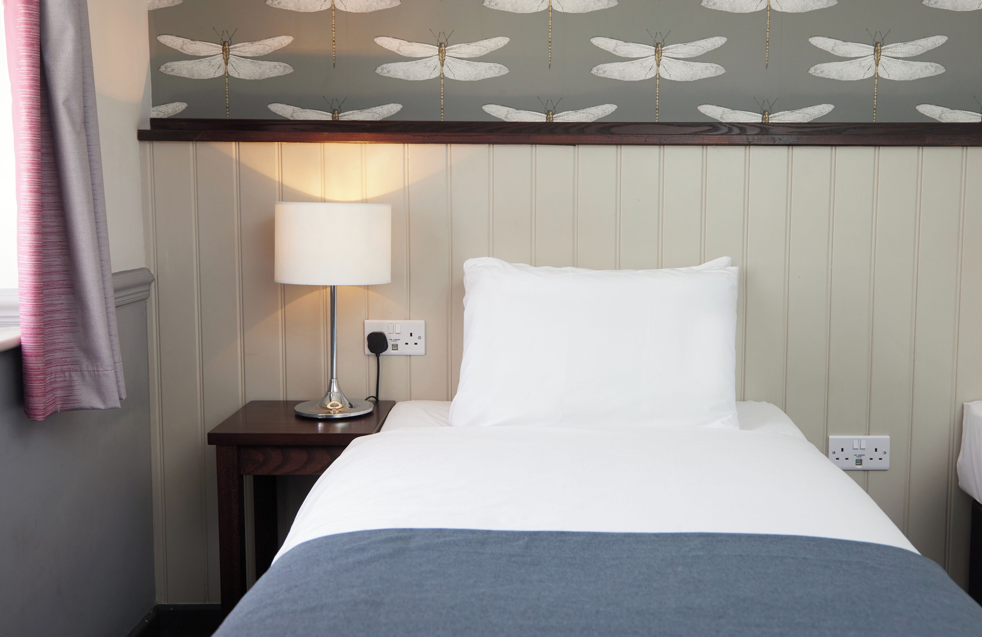 A neatly made bed with a white pillow and a blue blanket, a bedside lamp, and a wall with a dragonfly pattern.