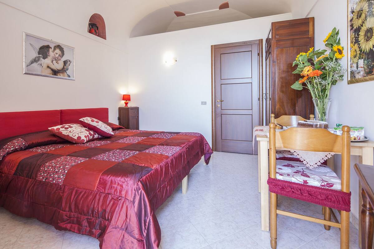 A bedroom with a red bed, a wooden chair, a desk with a vase of flowers, and a door.